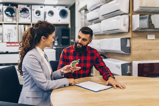 A man and saleswoman sitting at a table with a clipboard with appliances and HVAC systems for sale in the background.